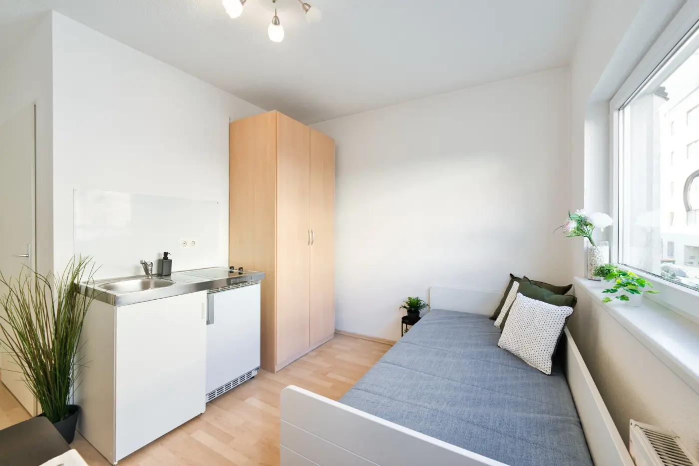 Studio bei Floren: Bett am Fenster, Kleiderschrank und Küchenzeile in einem hellen Raum